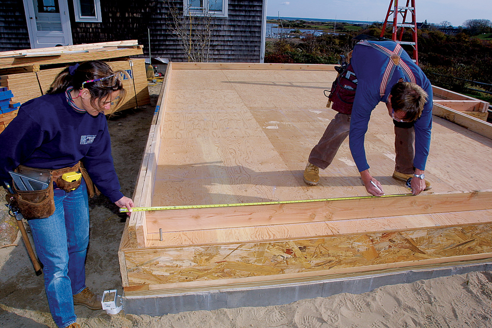 Stud layout is always taken from the same two walls. One crew member holds the tape at wall offset while the other marks the stud position.