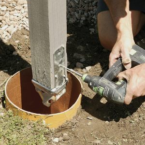 Attach the hardware, then pour. After locating and backfilling the tube, fasten the galvanized post base to the existing post.