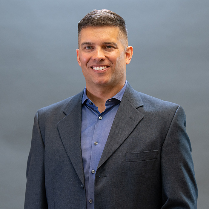 Headshot of Brendan Knapmam in a dark gray suit against a blue background