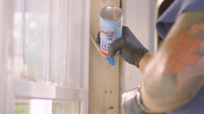 A close up of a hand wearing black rubber gloves using a bottle of Loctite TITE FOAM Window & Door to air seal the area.