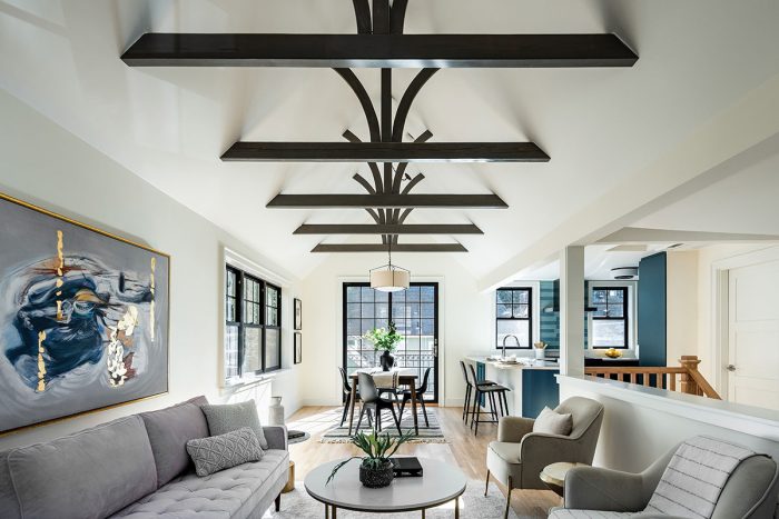 Photo of a vaulted ceiling featuring wooden beams spanning across the middle with white ceilings and walls, grey furniture, and a blue painting.