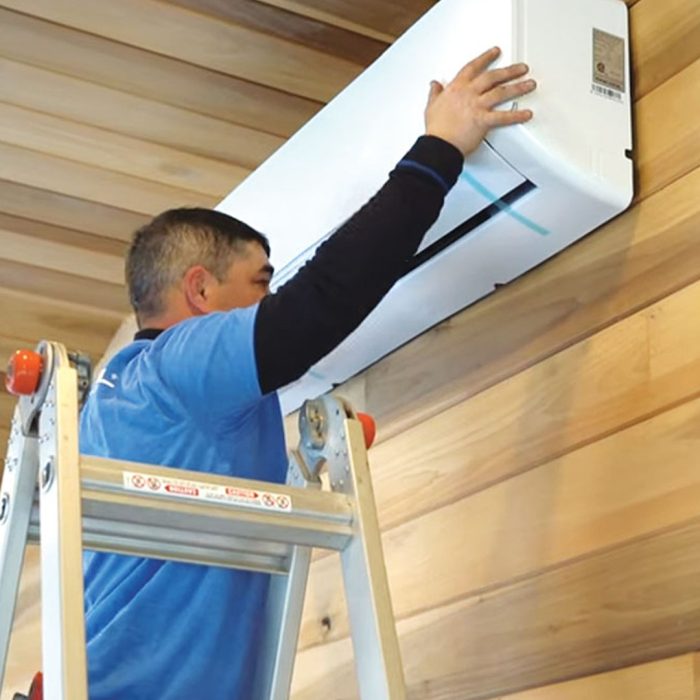 Man in a blue shirt installing a minisplit in a room with wood board walls and ceilings.